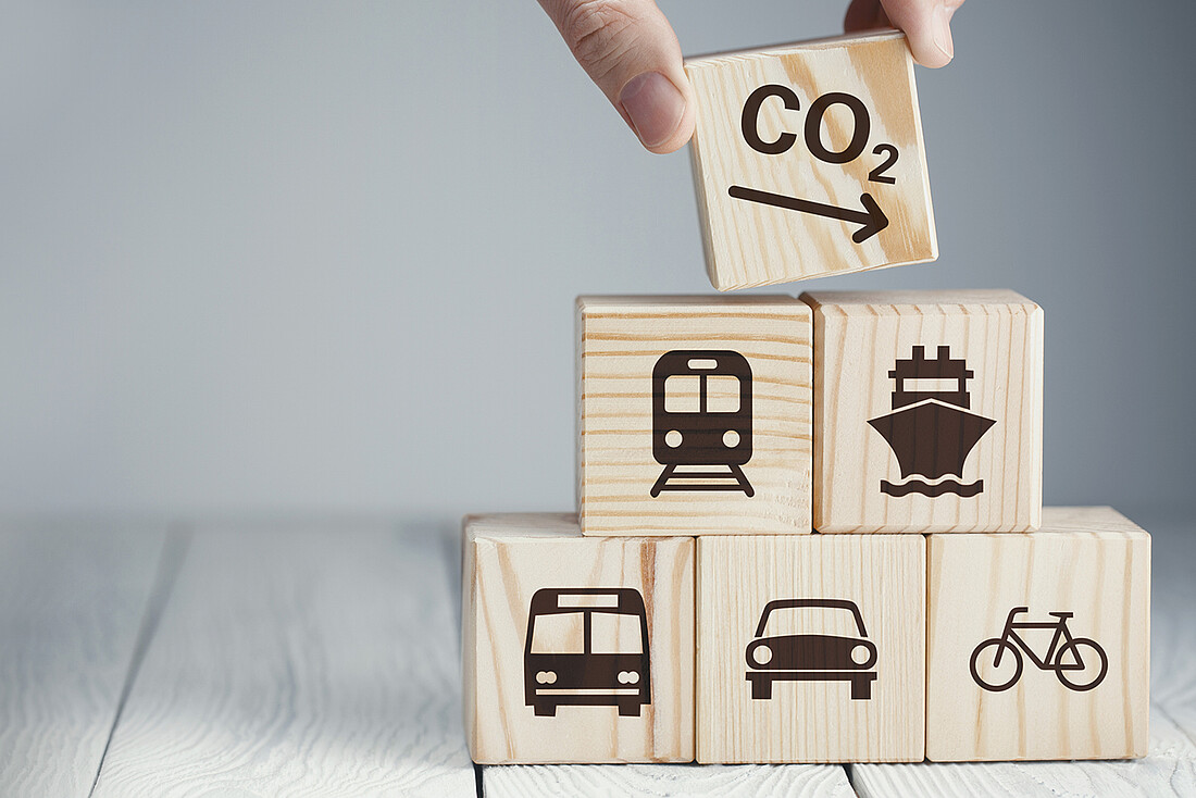 Fünf aufeinandergestapelte Würfel aus Holz mit Symbolen, die den Verlehrssektor repräsentieren (Bus, PKW, Fahrrad, Zug, Schiff) und ein Würfel mit einem Symbol für sinkende CO2-Emissionen